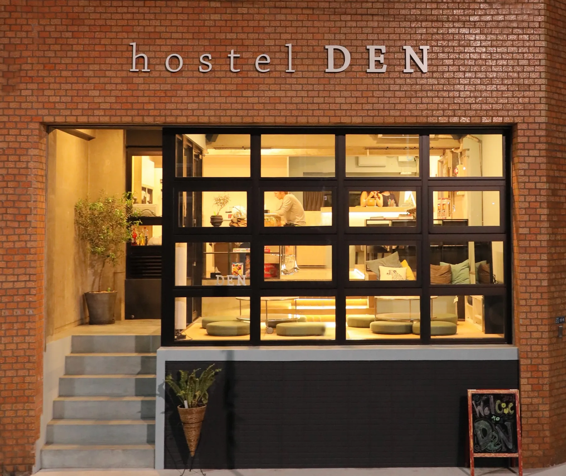 The red-brick front of hostel DEN at night, with the lounge lit up behind a large black-framed window